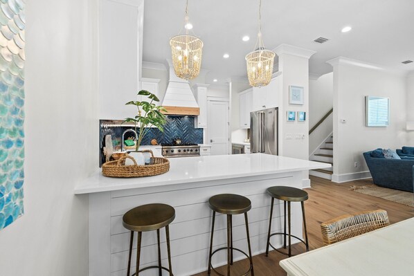 A modern kitchen with a white island, bar stools, and hanging chandeliers.