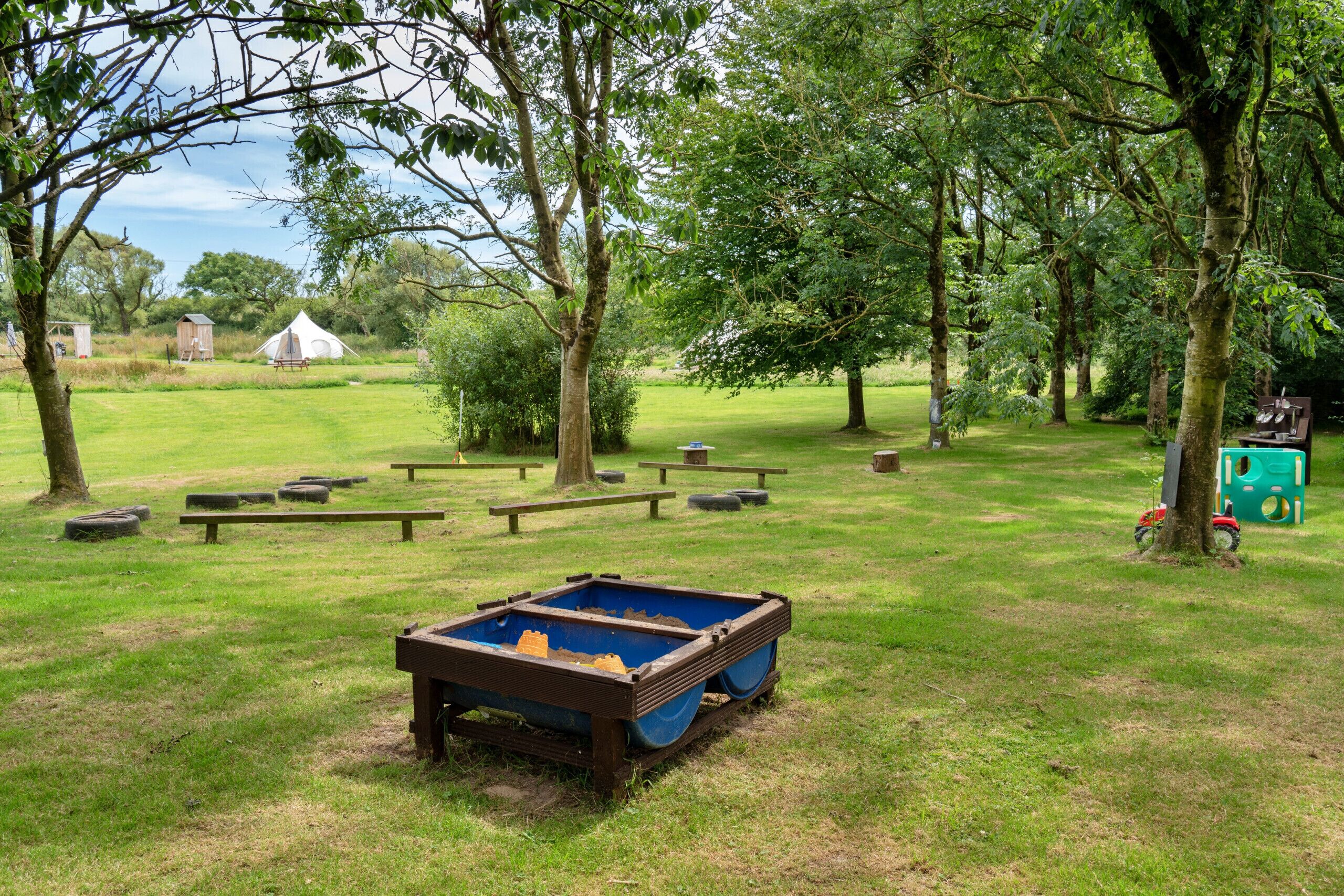 Un terrain de camping avec des tables de pique-nique, un conteneur bleu et une tente, situé dans une zone herbeuse bordée d’arbres.