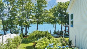 Vista de un lago con una casa blanca, una terraza de madera y un jardín con hortensias.