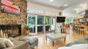 Un salón espacioso con chimenea de piedra, un televisor grande y un comedor con sillas de madera, con una cocina al fondo.