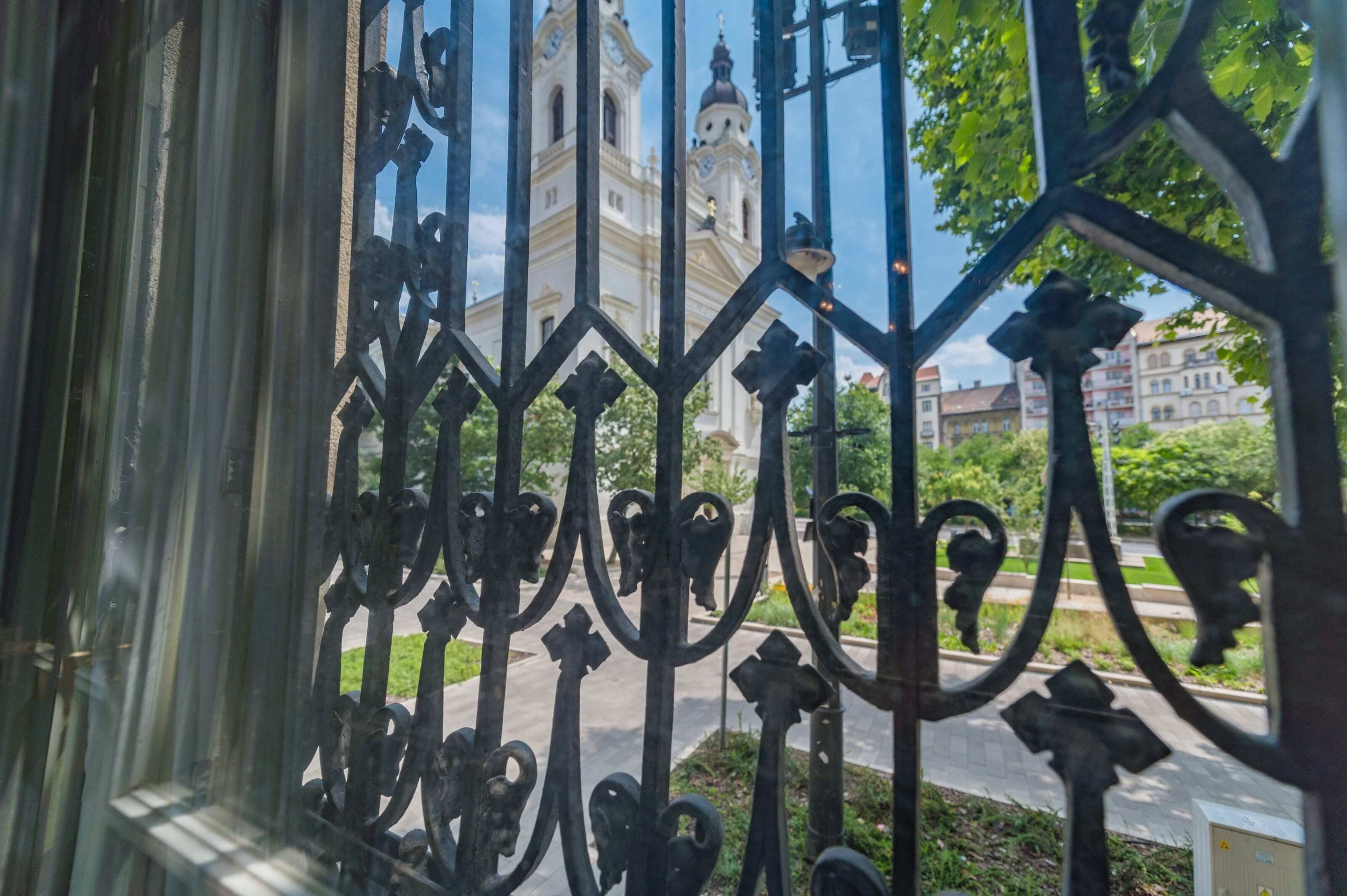A wrought iron gate with intricate designs, leading to a street with a historic building in the background.