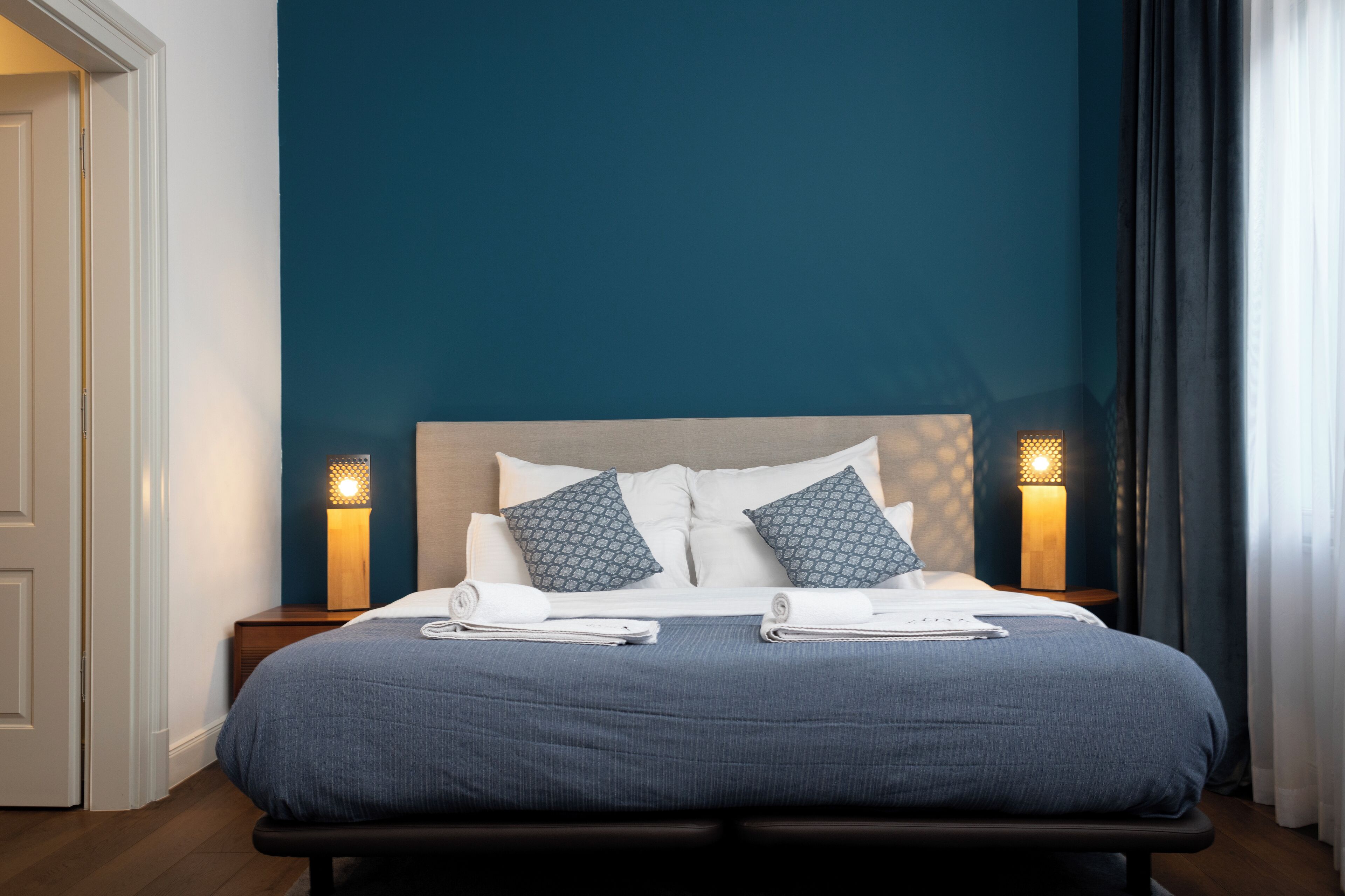 A neatly made bed with a blue bedspread, two patterned pillows, and two bedside lamps.