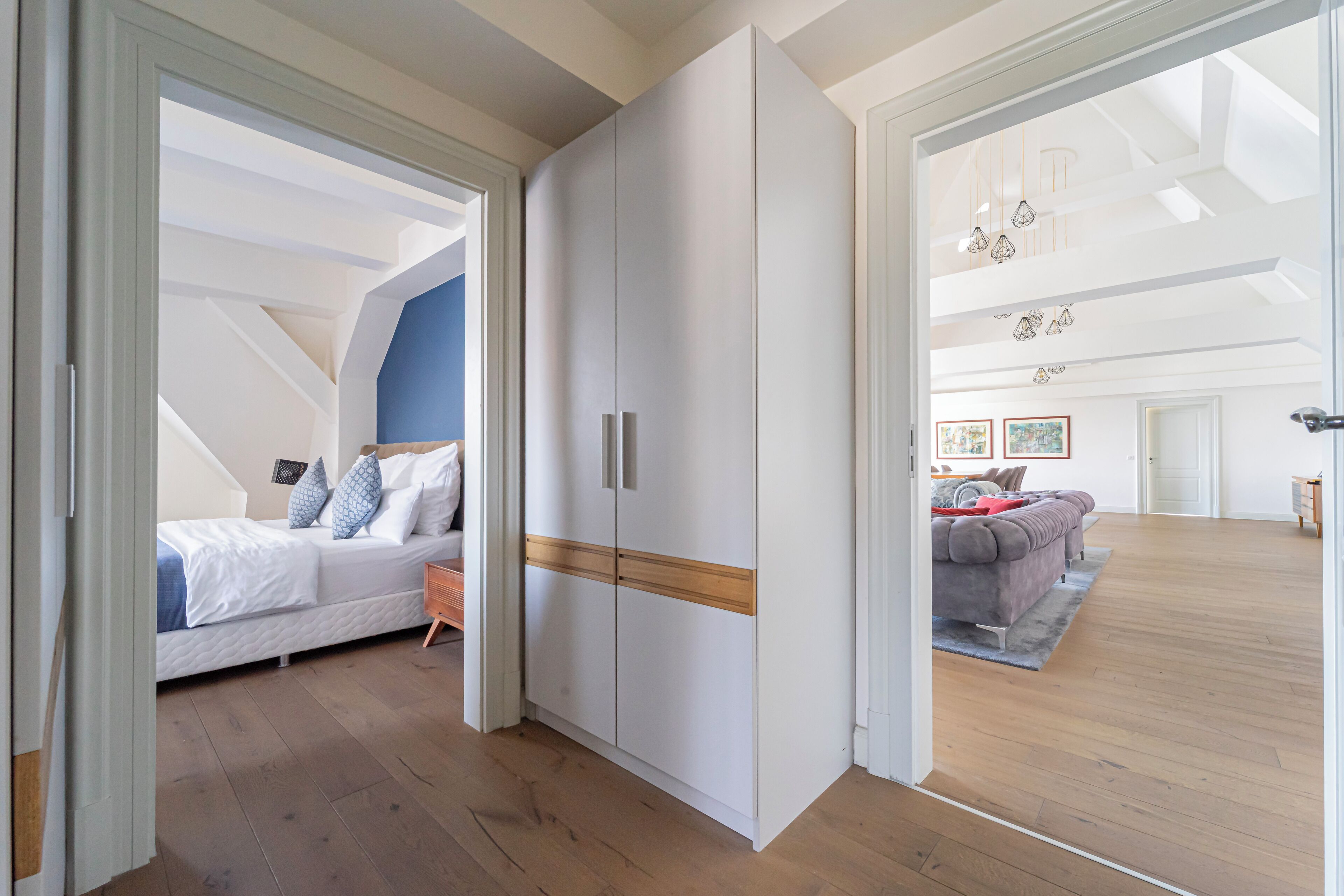 Une pièce spacieuse avec une grande armoire, un lit avec une literie bleue et blanche, et une vue sur une autre pièce où se trouvent un canapé et un tapis.