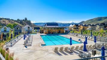A swimming pool with lounge chairs and umbrellas, surrounded by modern buildings with a mountainous backdrop.