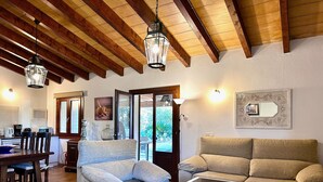 A cozy living room with a sofa, armchair, and coffee table, wooden beams on the ceiling, and a dining area with chairs and a table. - Holidayrental Majorca - Can Puig (Pollensa / Pollença)