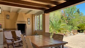 A covered patio with a wooden table and chairs, a fireplace, and a view of a garden with various plants. - Holidayrental Majorca - Can Puig (Pollensa / Pollença)