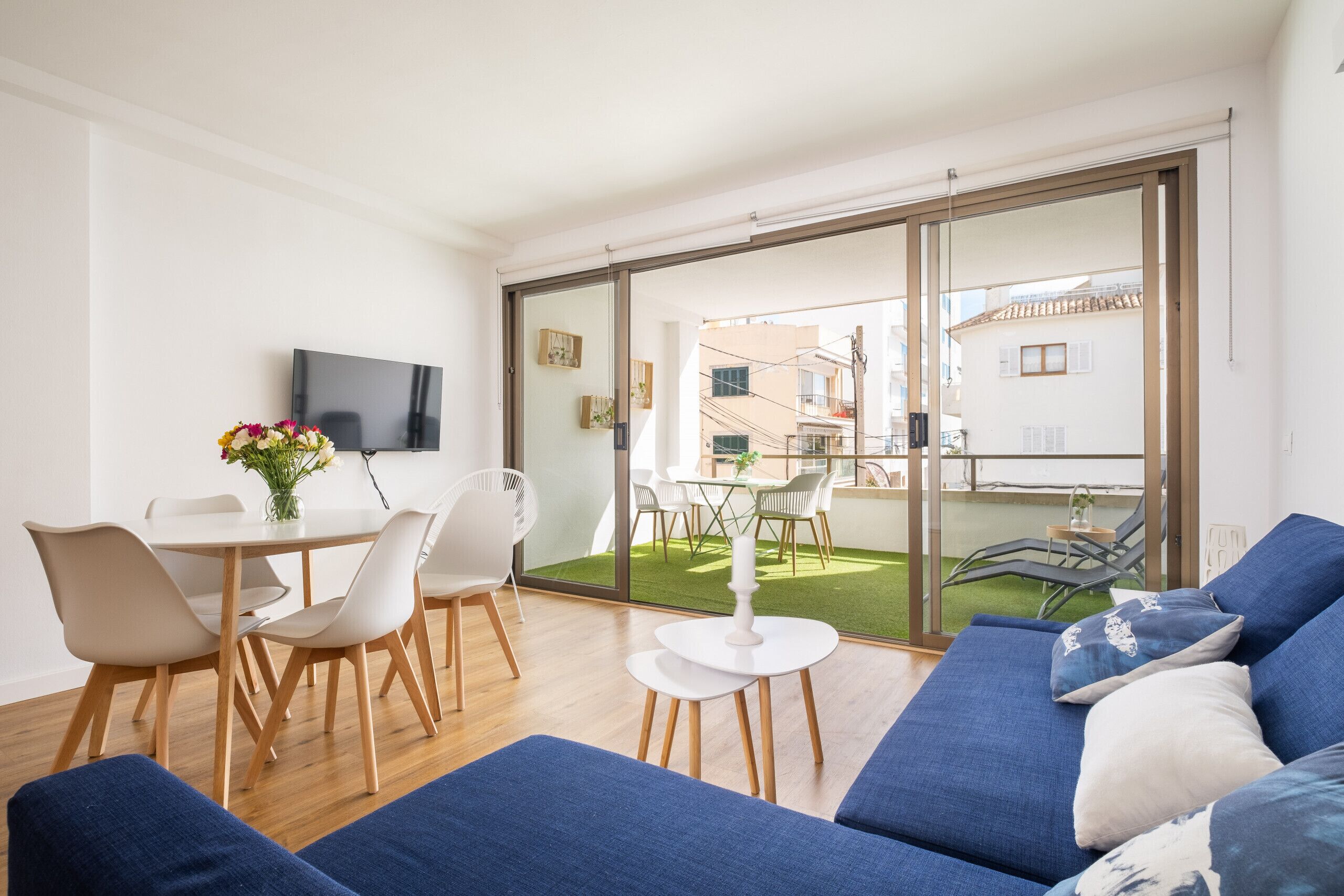 A modern living room with a sofa, dining table, and chairs. There is a TV mounted on the wall and a balcony with outdoor furniture.
