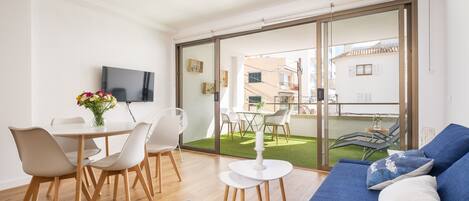 A modern living room with a sofa, dining table, and chairs. There is a TV mounted on the wall and a balcony with outdoor furniture.