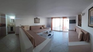 Ein geräumiges Wohnzimmer mit einer Sofagarnitur, einem Couchtisch und einem im Hintergrund sichtbaren Küchenbereich.