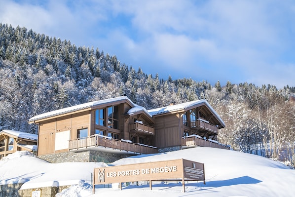 Una cabaña de madera con balcones, un cartel que dice "Les Portes de Megève" y un paisaje montañoso nevado.