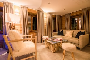Una sala acogedora con piso de madera, un sillón blanco mullido, una mesa de centro de vidrio, un sofá beige con almohadones negros y cortinas en las ventanas.