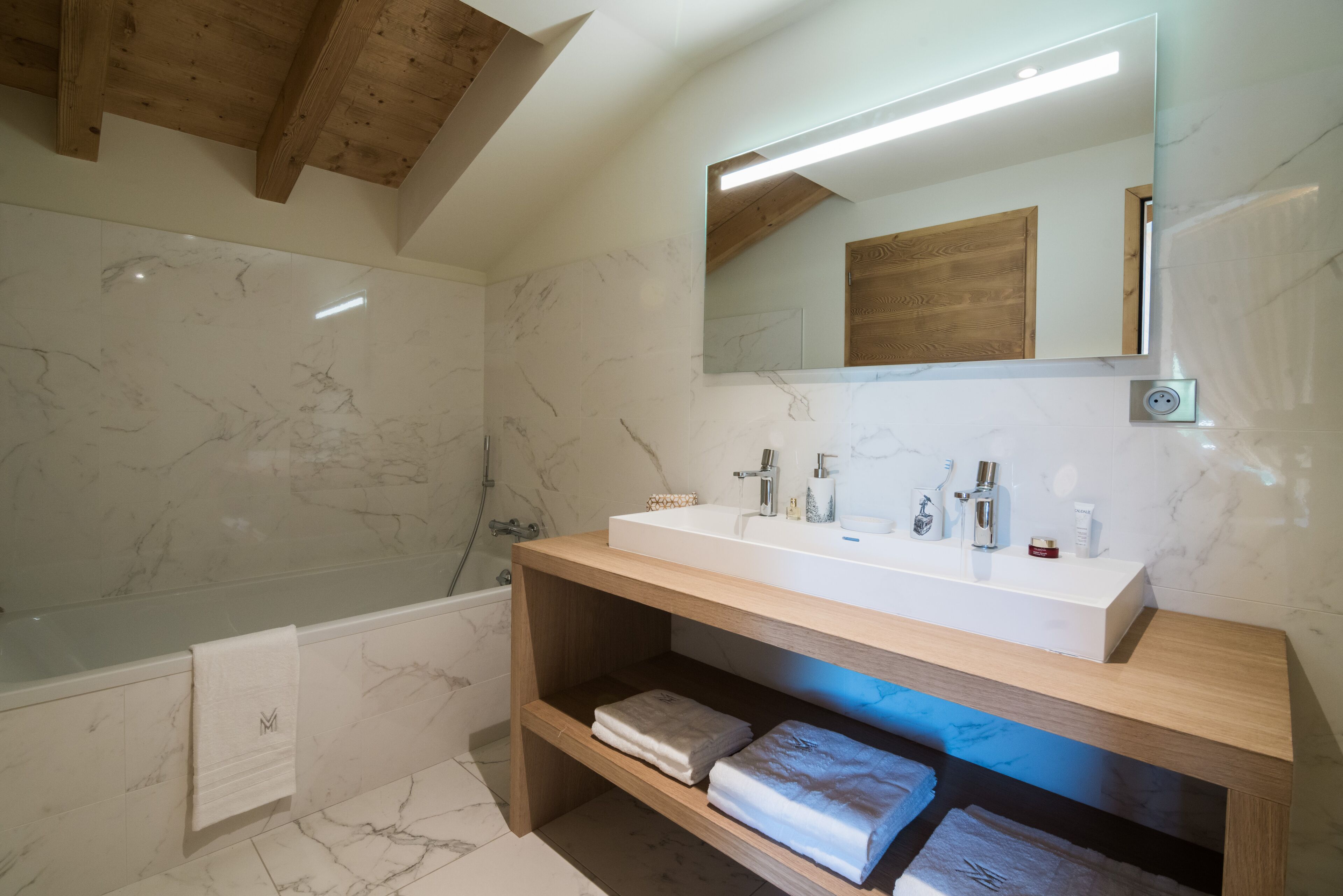 Een moderne badkamer met een houten wastafelmeubel, marmeren tegels, een grote spiegel en een bad.