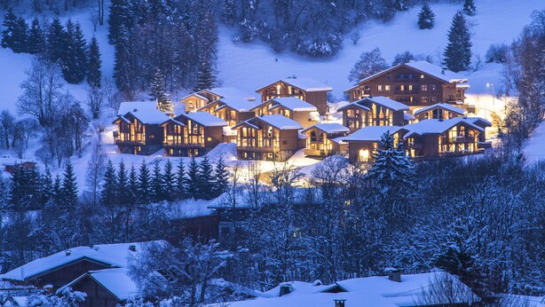 Un gruppo di baite in legno con tetti imbiancati dalla neve, circondate da alberi, in zona montana.
