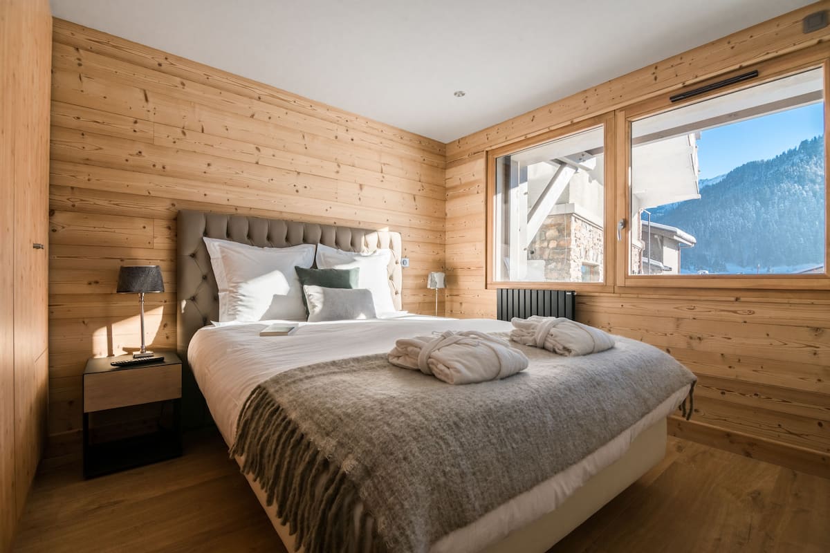Une chambre à coucher aux murs lambrissés, avec un grand lit, une table de chevet, une lampe et une vue sur une montagne par la fenêtre.