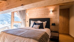 Una camera da letto con soffitto in legno, un letto grande con coperta bianco-marrone, un comodino con una lampada e una finestra con tende.