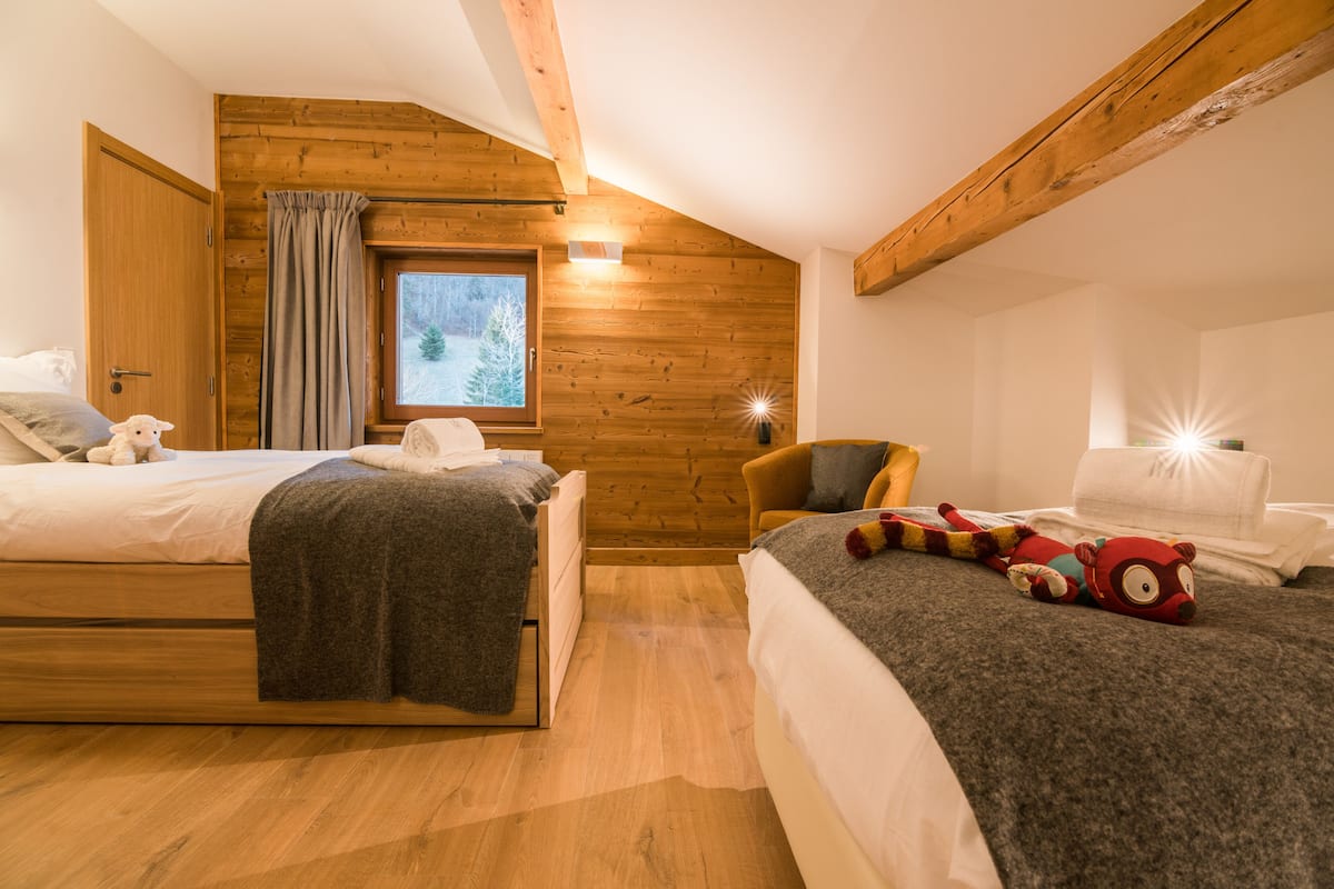 Une chambre confortable avec deux lits, des murs en bois, une fenêtre donnant sur les arbres et un jouet en peluche sur l’un des lits.