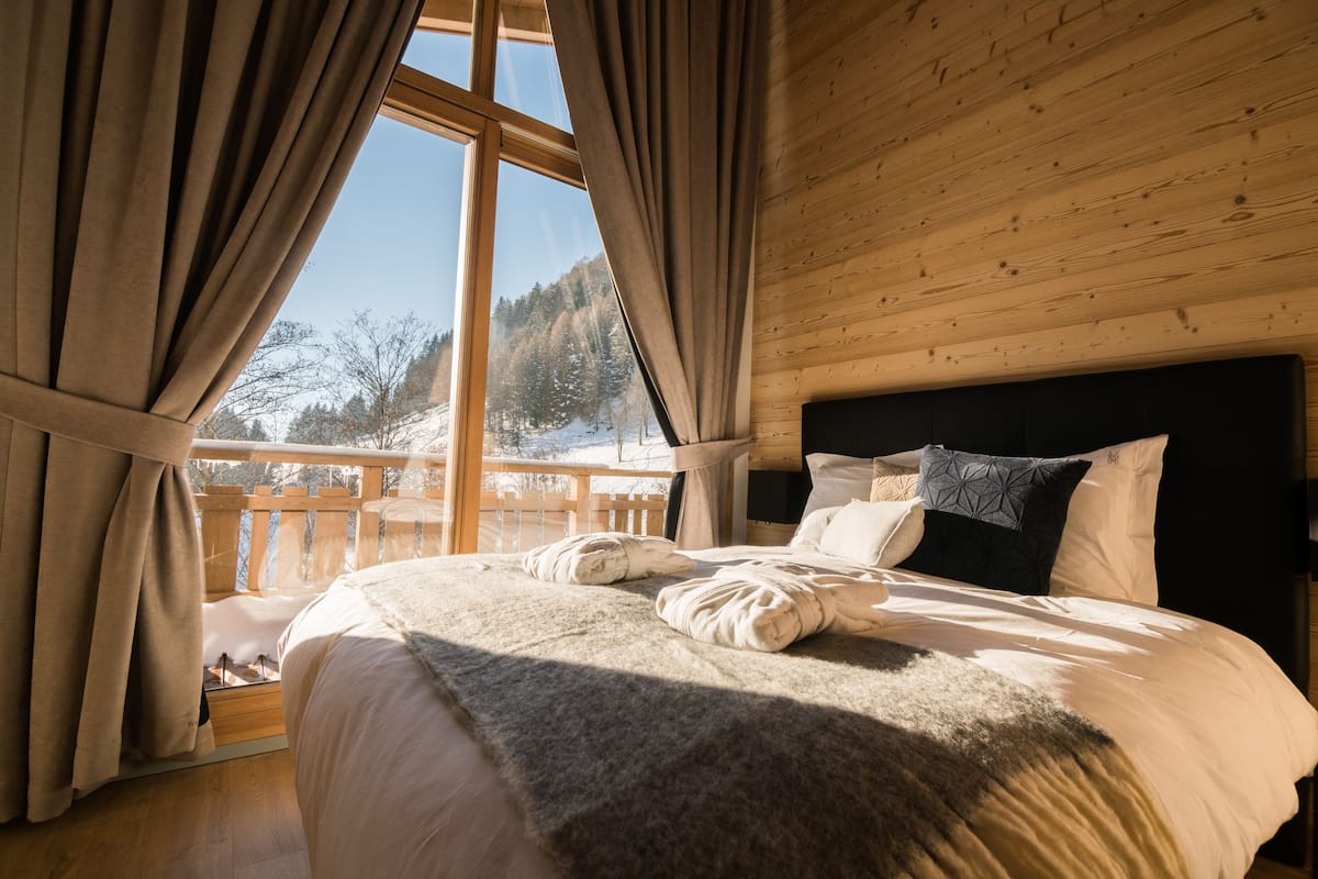 Une chambre avec une grande fenêtre, un plafond en bois et un lit recouvert d’une literie blanche et d’un plaid gris.