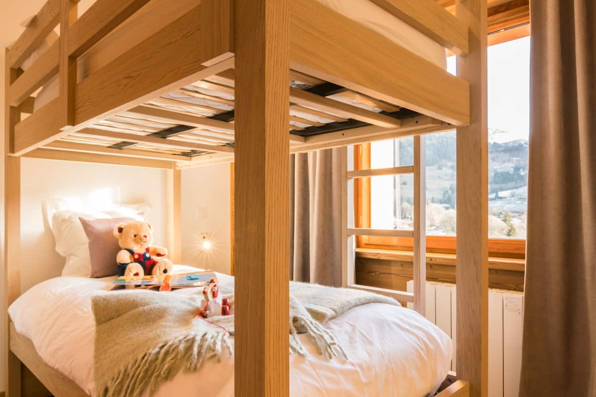 Un lit superposé avec une structure en bois, une table de chevet avec une lampe, un ours en peluche et une fenêtre avec des rideaux.