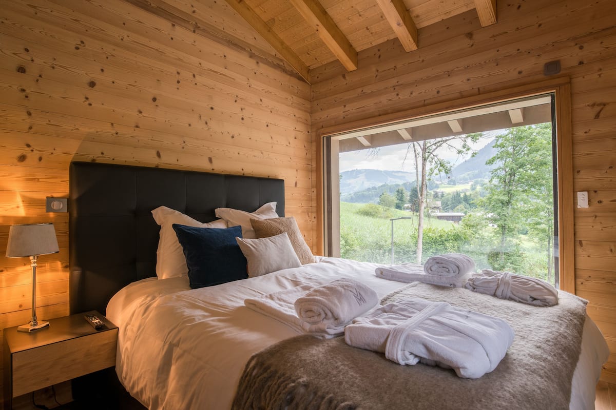 Une chambre aux murs lambrissés, dotée d’une grande fenêtre, d’un lit recouvert de draps blancs et de serviettes posées sur le lit.