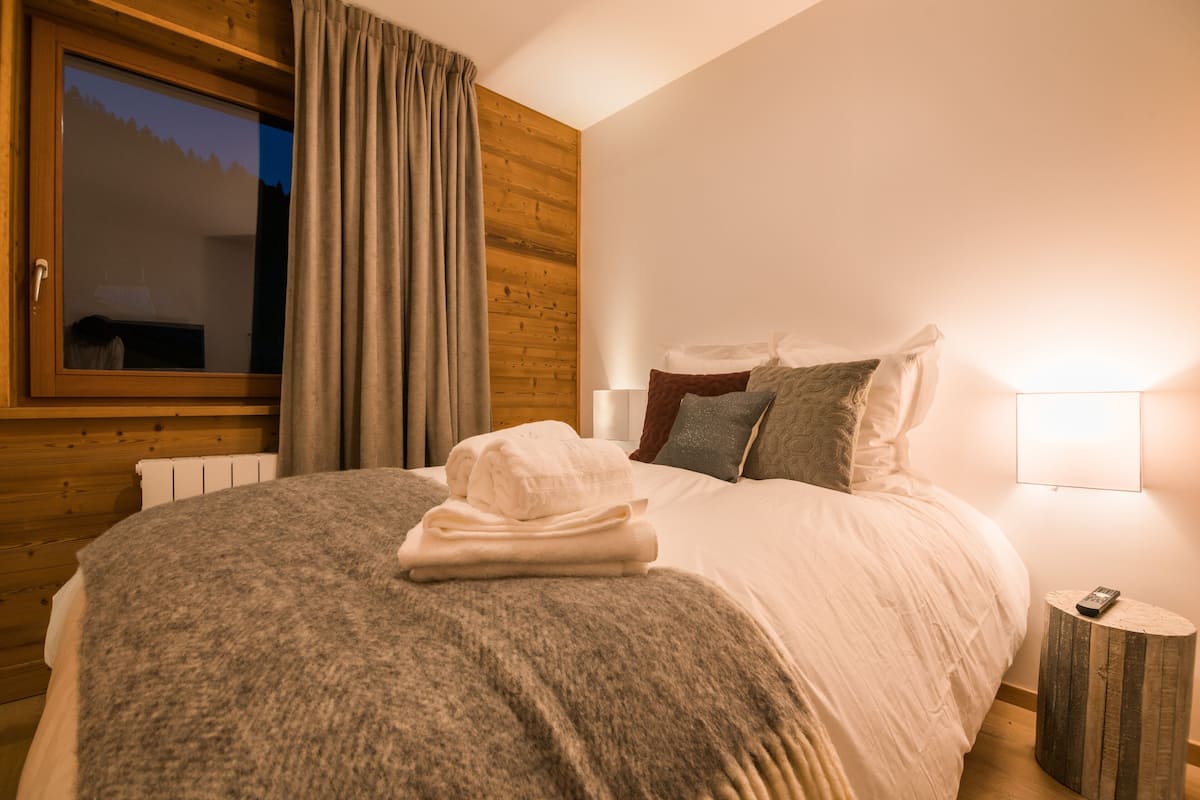 Un lit bien fait, avec une couverture grise et des serviettes pliées, une lampe de chevet et une tête de lit en bois.