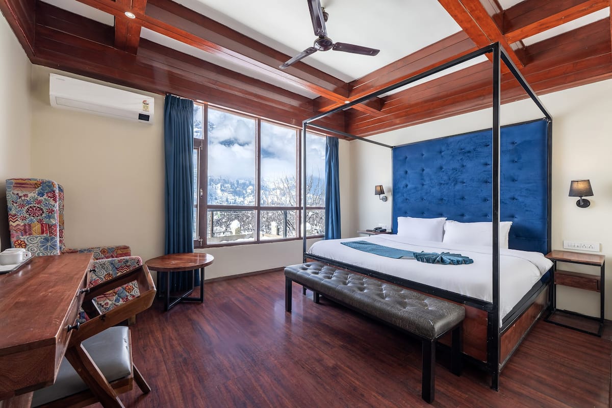 Una cama con dosel, cabecera capitonada, un banco de madera, una mesita redonda pequeña y una silla colorida en una habitación con suelo de madera y un ventanal grande.