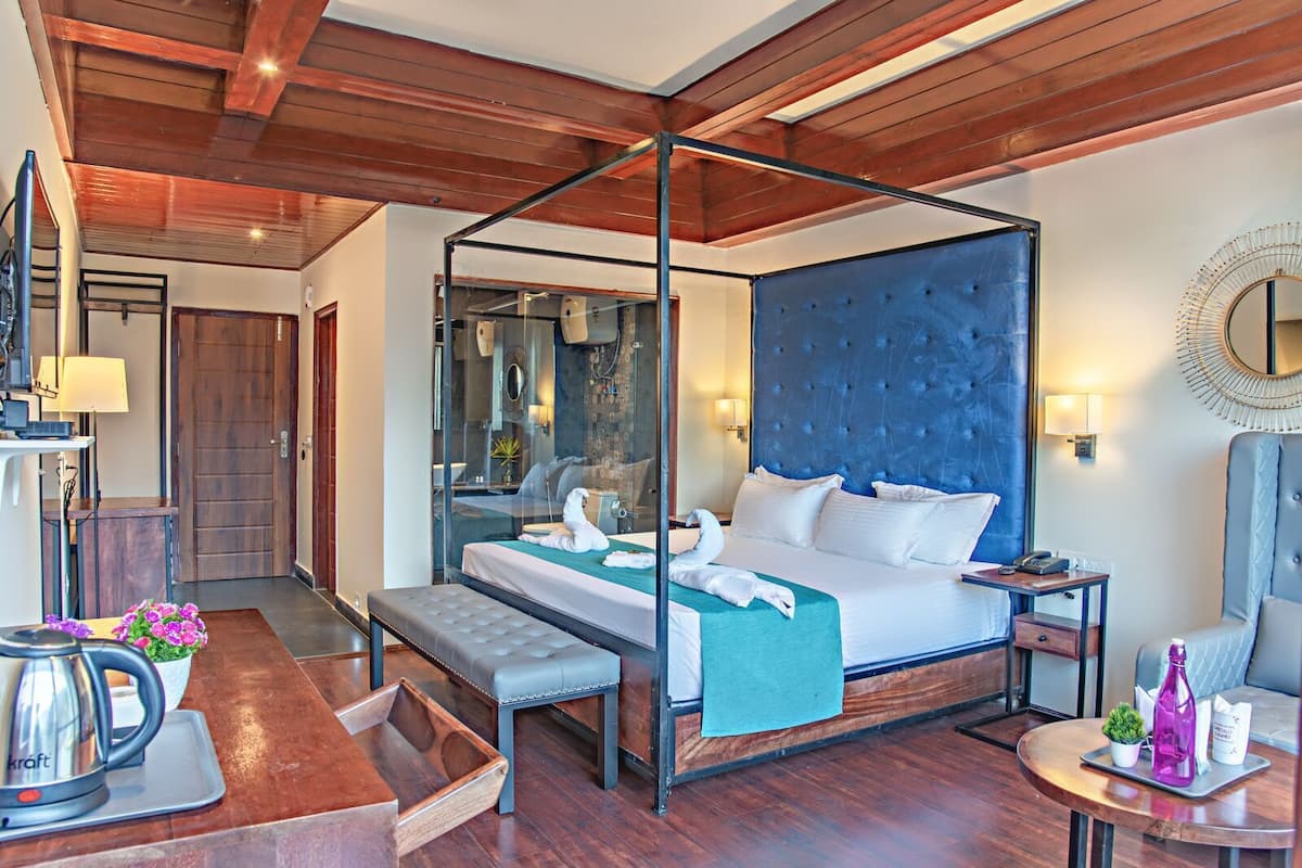 Habitación de hotel con cama con dosel, un banco, una mesita de noche, un espejo y una pequeña mesa con una botella y una planta.