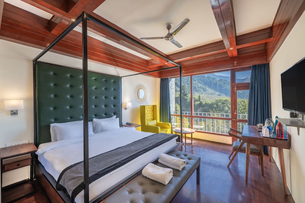 Una cama con dosel y un cabecero capitonado, techo de madera con vigas a la vista, balcón con vistas a montañas, un sillón amarillo y un televisor sobre una mesa de madera.