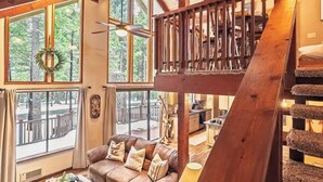 A cozy cabin interior with a wooden staircase, a living room with a sofa and coffee table, and large windows offering a view of the surrounding forest. - Evergreen Pinecone Cottage • Hot Tub • Pool Access • Espresso • A/C (Dorrington)
