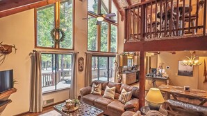 A spacious living room with a large brown leather sofa, a coffee table, and a fireplace. There are large windows providing natural light and a view of the outdoors. - Evergreen Pinecone Cottage • Hot Tub • Pool Access • Espresso • A/C (Dorrington)