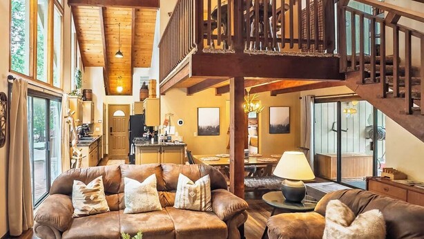 A spacious living room with a high ceiling, wooden beams, and a cozy fireplace. There is a dining area with a table and chairs, and a staircase leading to an upper level. - Evergreen Pinecone Cottage • Hot Tub • Pool Access • Espresso • A/C (Dorrington)