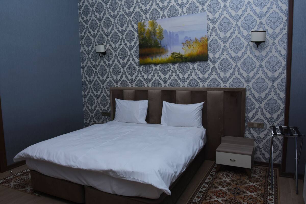 A neatly made bed with white linens, a bedside table with a drawer, a wall-mounted lamp, and a painting of a landscape.