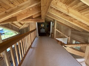 Cozy attic room with a high ceiling, carpeted floor, and wooden railing. Features a bed, bedside table, and mirror.