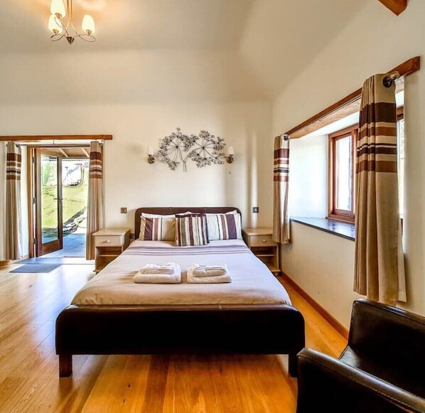 Une chambre à coucher avec un grand lit, un plancher en bois, un lustre, une fenêtre avec des rideaux et une vue sur l’extérieur.