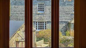 Vista a través de una ventana que muestra un edificio de piedra, un banco de madera y un jarrón con flores sobre una mesa.