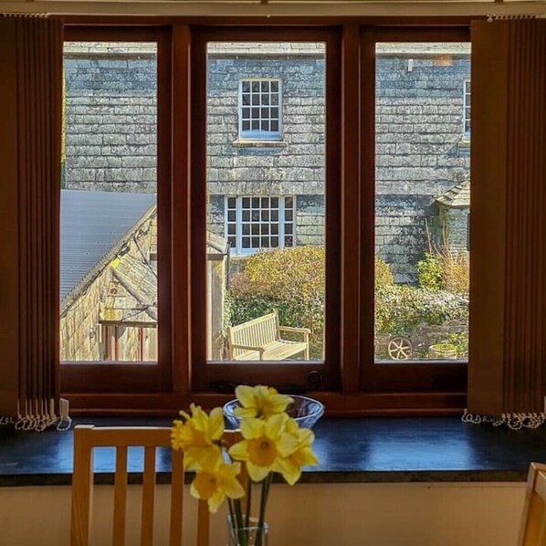 A view through a window with a stone building outside, a wooden bench, and a vase with flowers on a table.