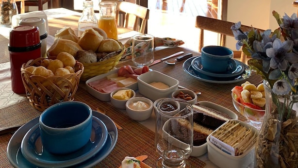 A breakfast table with bread, pastries, and beverages, a wooden ceiling, and a view of greenery outside. - Chalés Vista Linda (Santo Antonio do Pinhal)