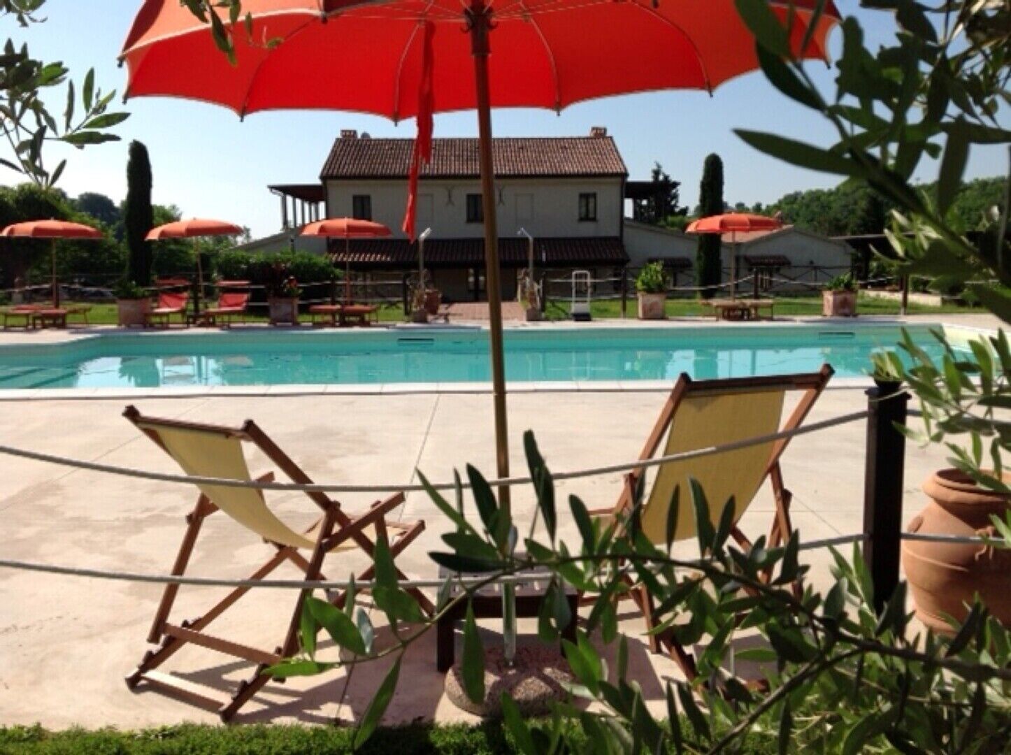 Area piscina con lettini e ombrelloni, una casa sullo sfondo e vegetazione circostante.