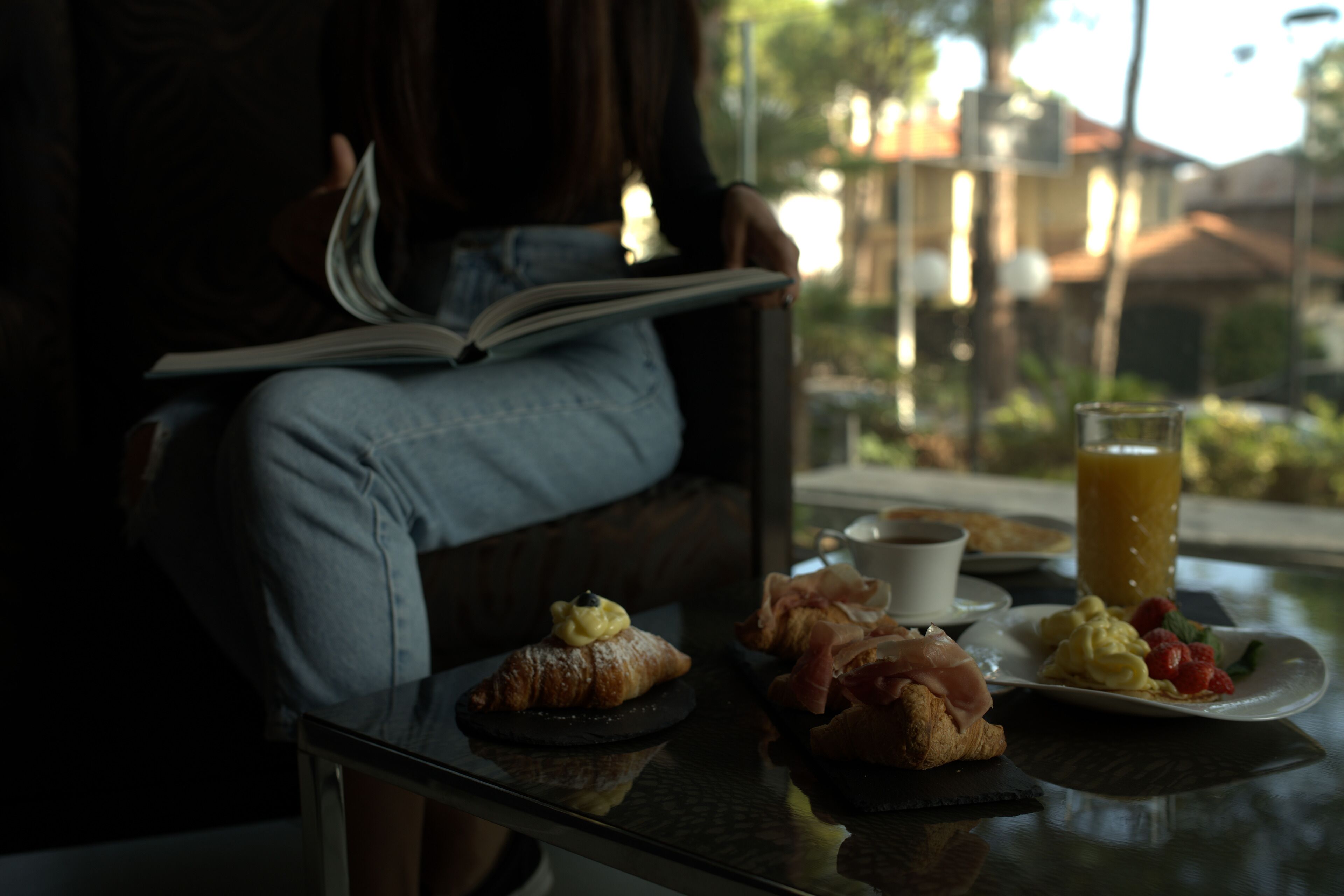 A person reading a book by a window with breakfast items on a table.
