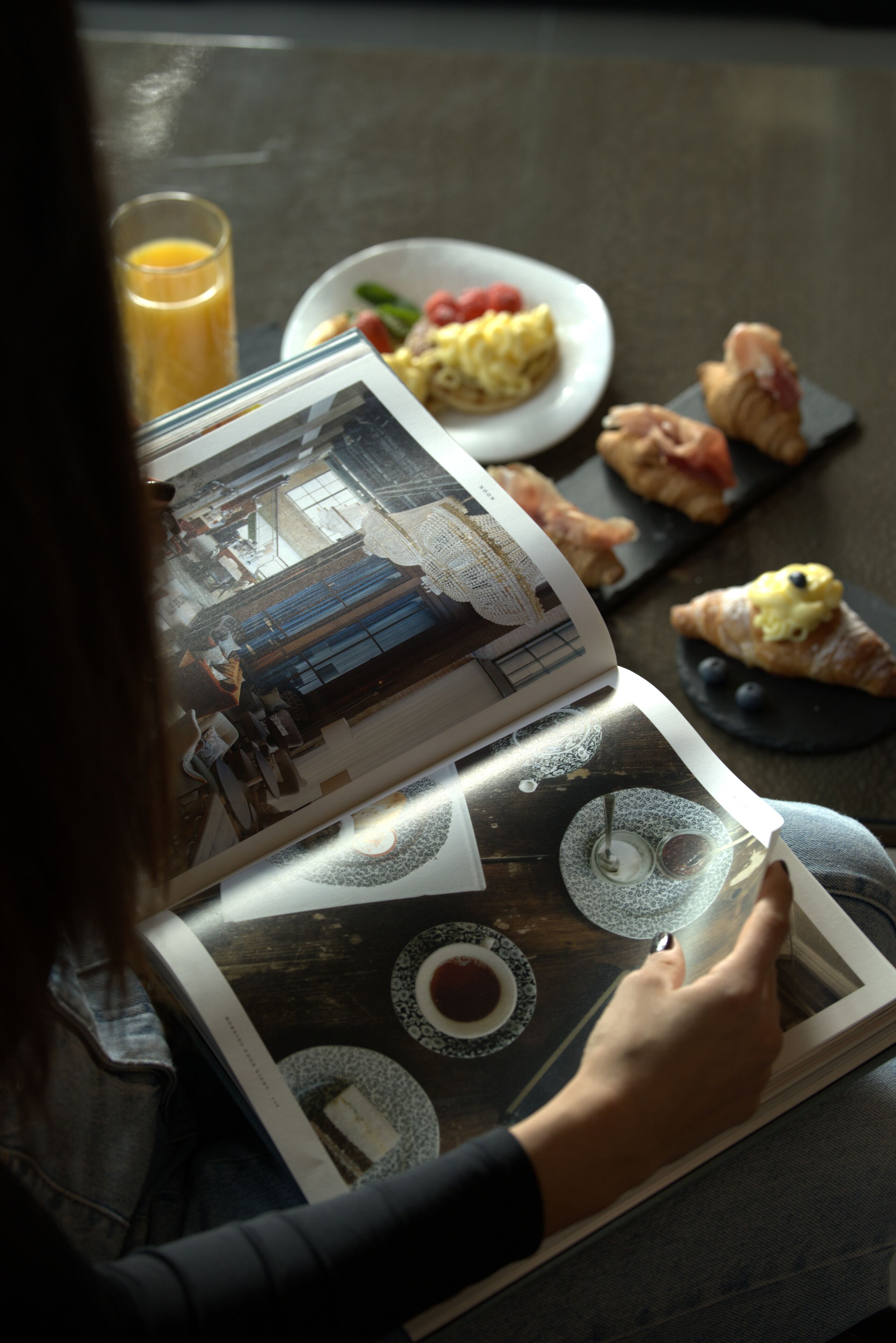 A person reading a magazine with a table in front of them, featuring food and a glass of orange juice.