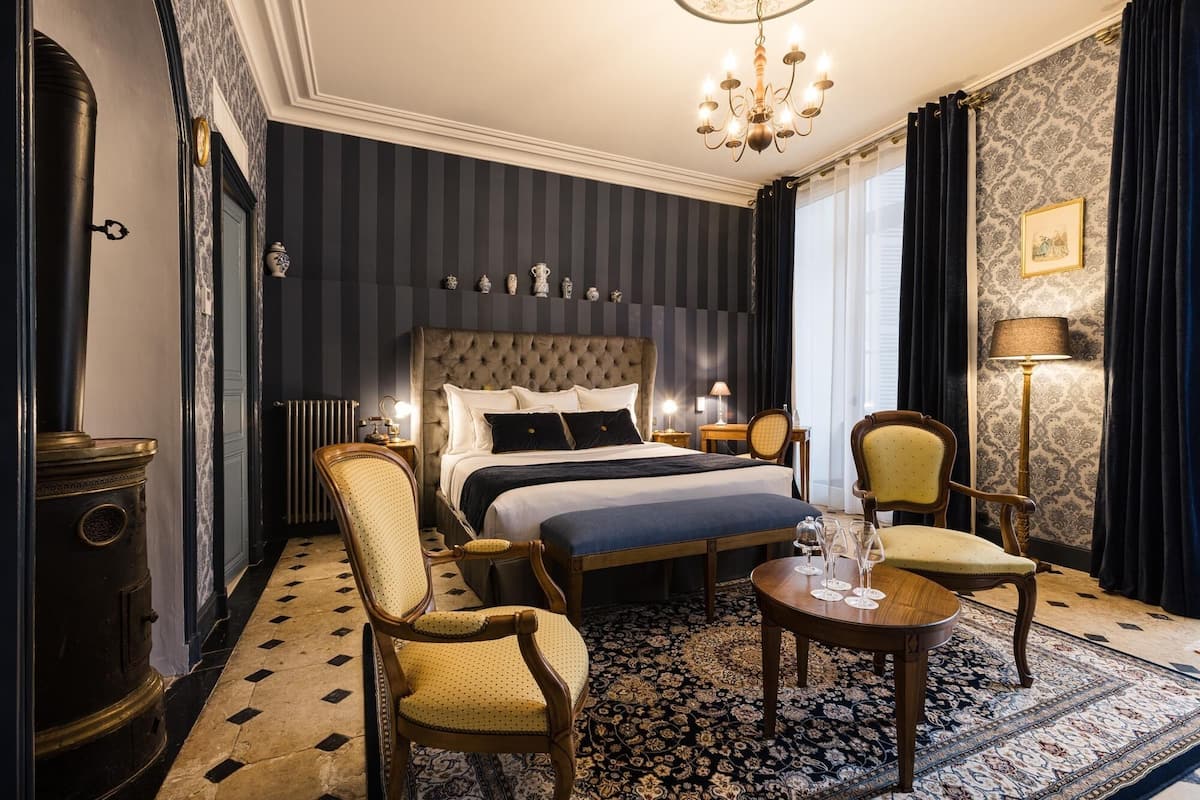 A hotel room with a large bed, two armchairs, a small table with glasses, a chandelier, and a patterned rug.