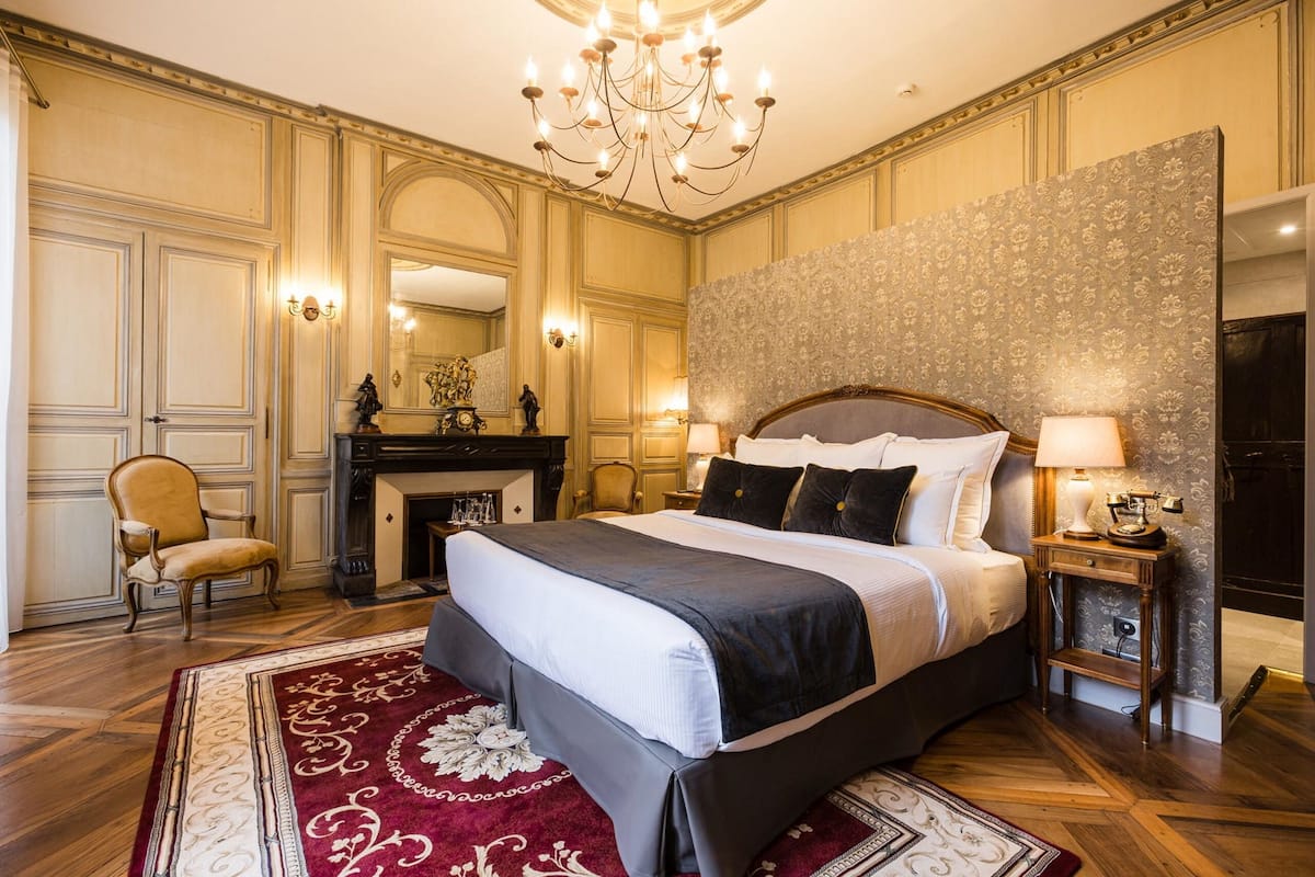 A luxurious bedroom with a large bed, a chandelier, a fireplace, and a classic decor style.