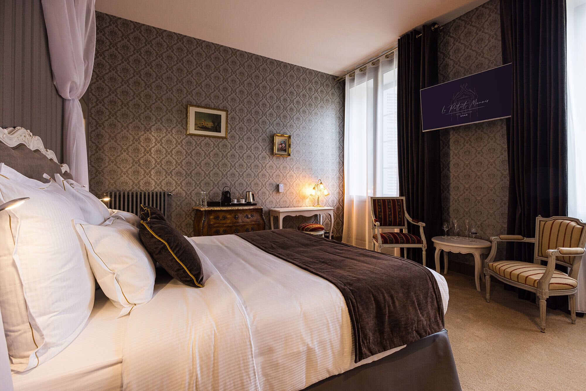 Une chambre d’hôtel avec un lit, une petite table avec une lampe, deux chaises et une grande fenêtre avec des rideaux.
