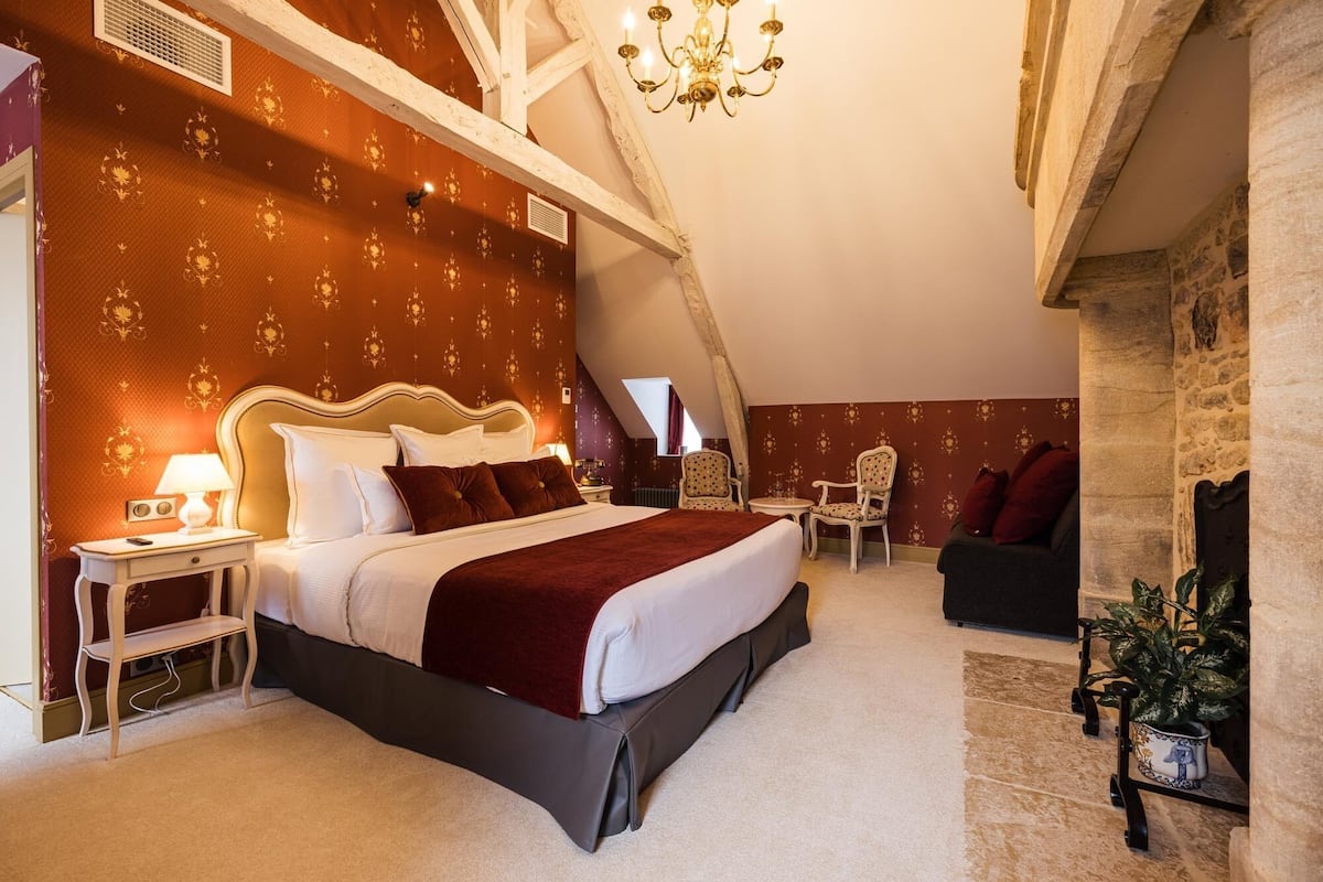 A bedroom with a large bed, a nightstand, a chair, a small table, and a fireplace.