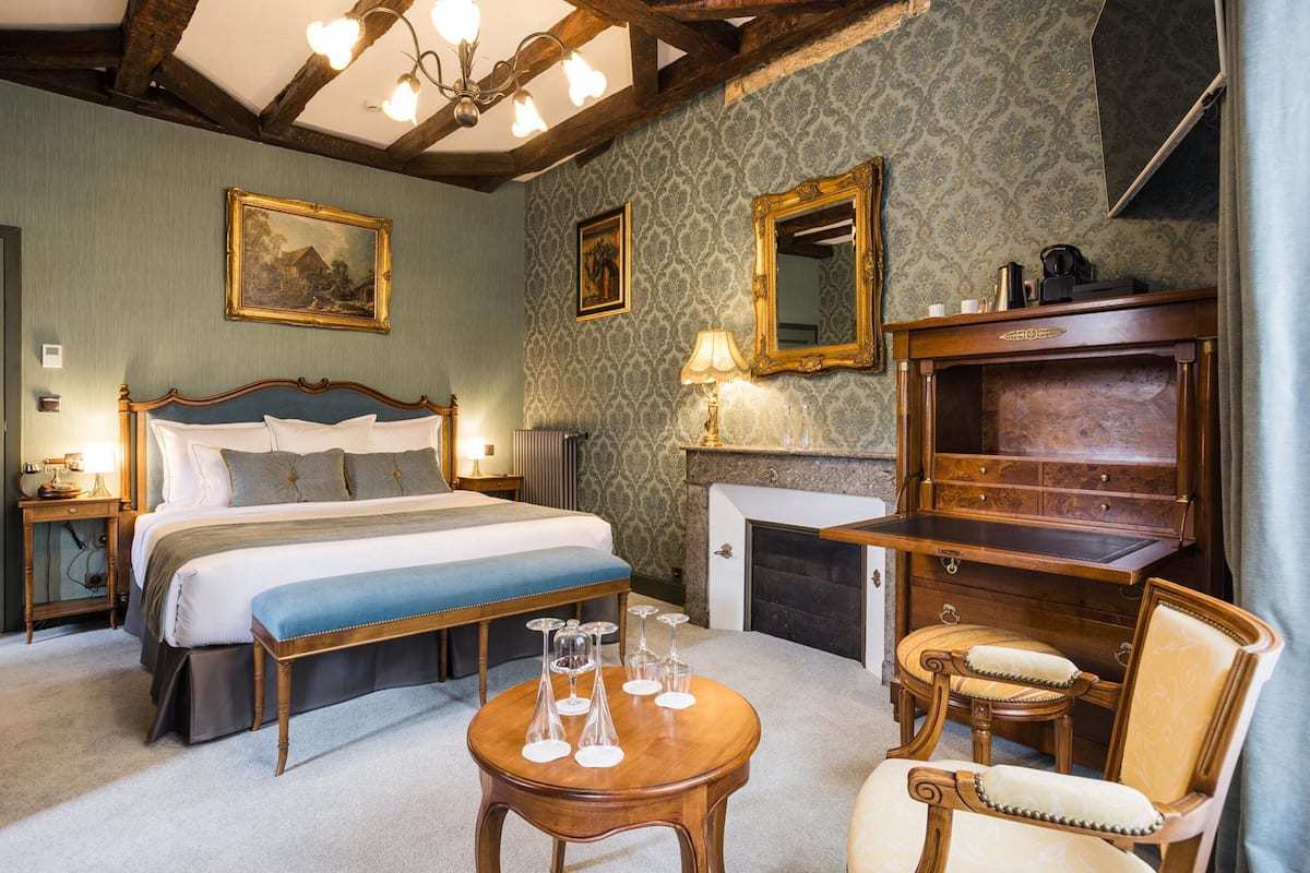 A hotel room with a large bed, a wooden desk, a chair, a fireplace, and a mirror on the wall.