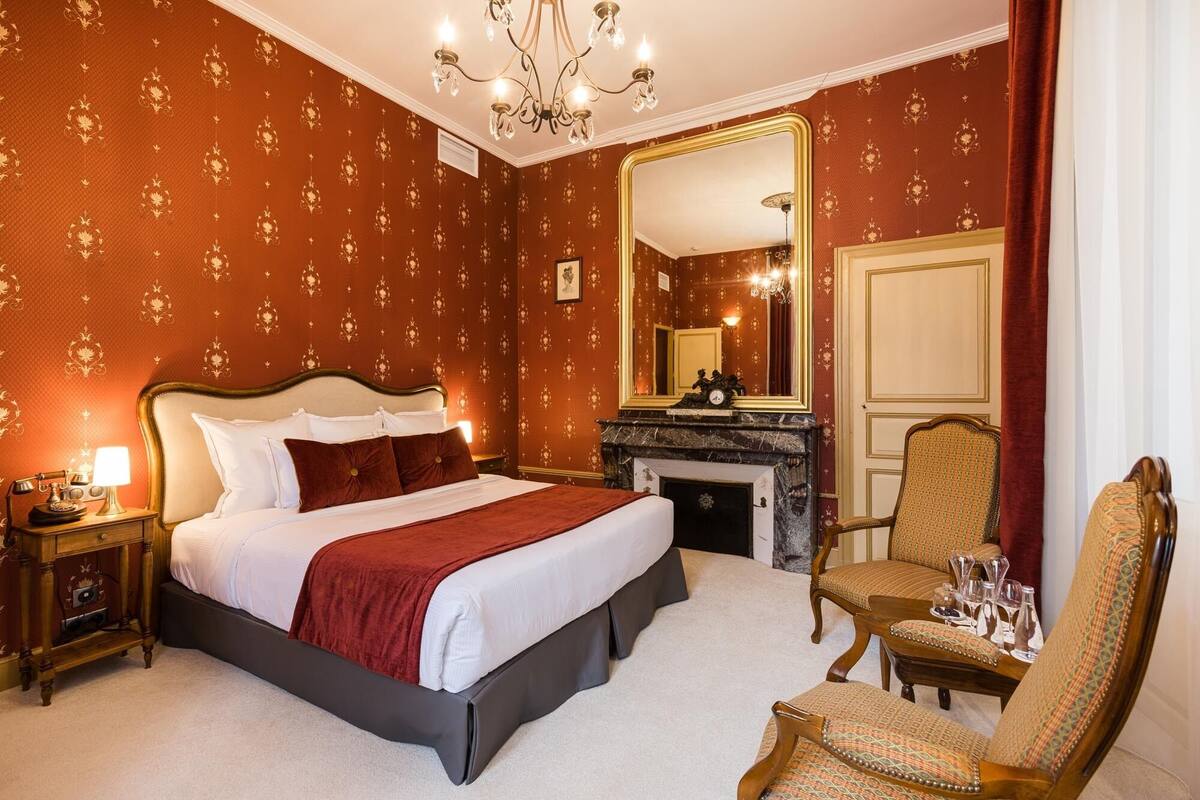 A hotel room with a large bed, a sitting area with a chair, a fireplace, and a large mirror.