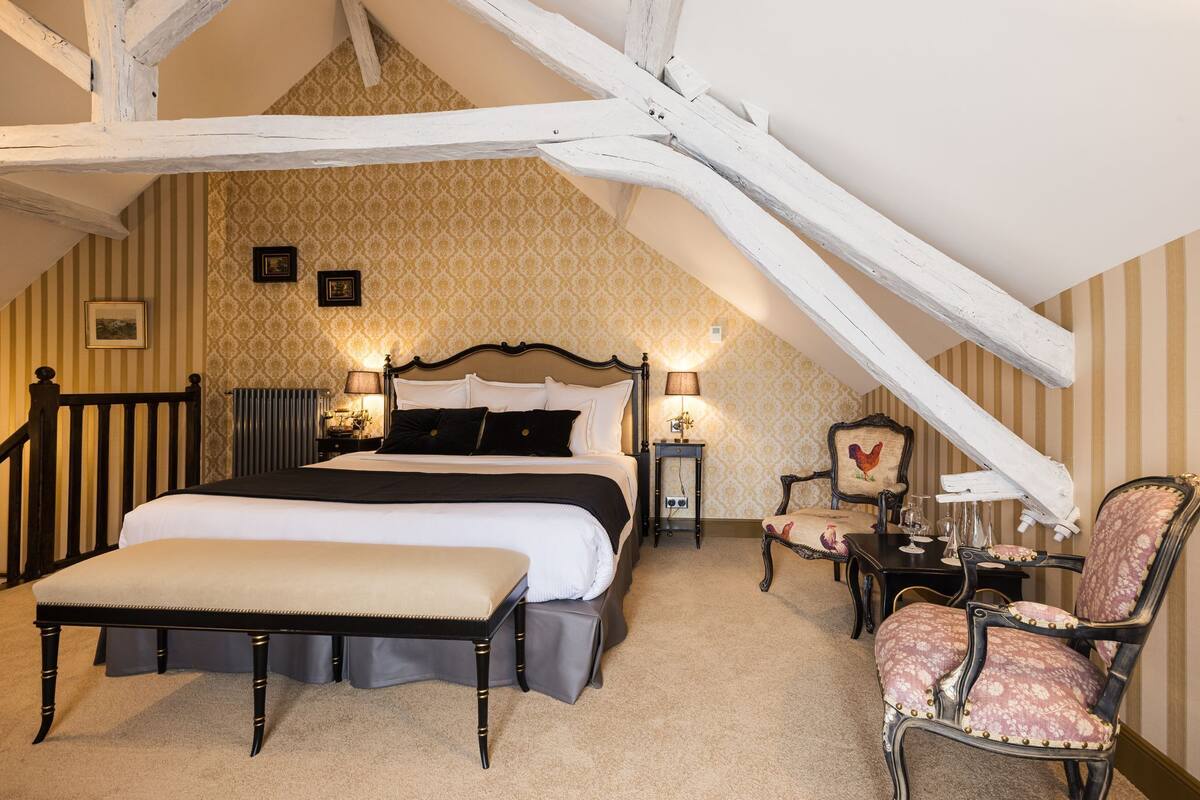 A bedroom with a large bed, a bedside table with a lamp, a chair, and a small table with a decorative item.