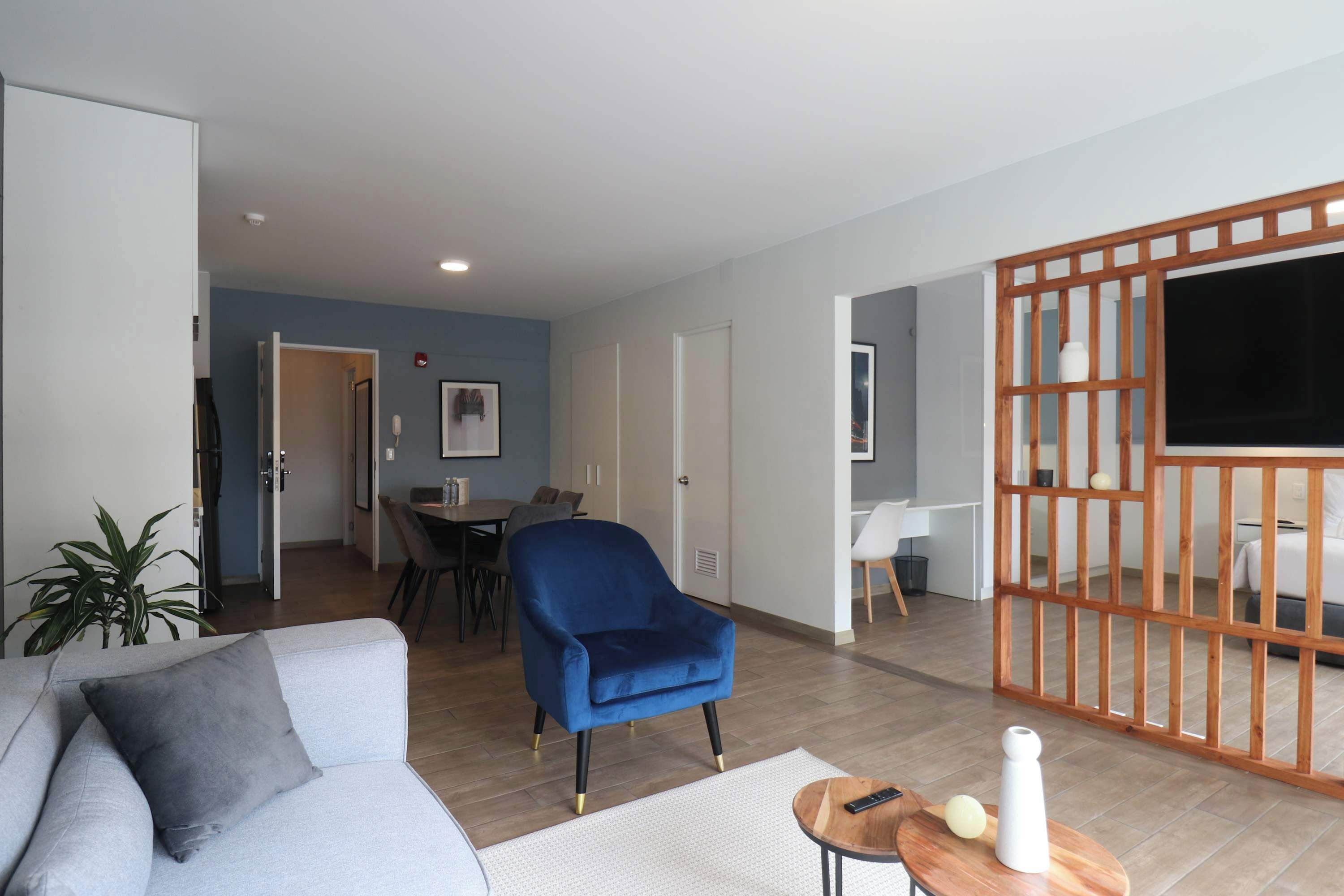 Un salon moderne avec un fauteuil bleu, un canapé gris et une table basse en bois. On aperçoit à l’arrière-plan un coin repas avec une table et des chaises.