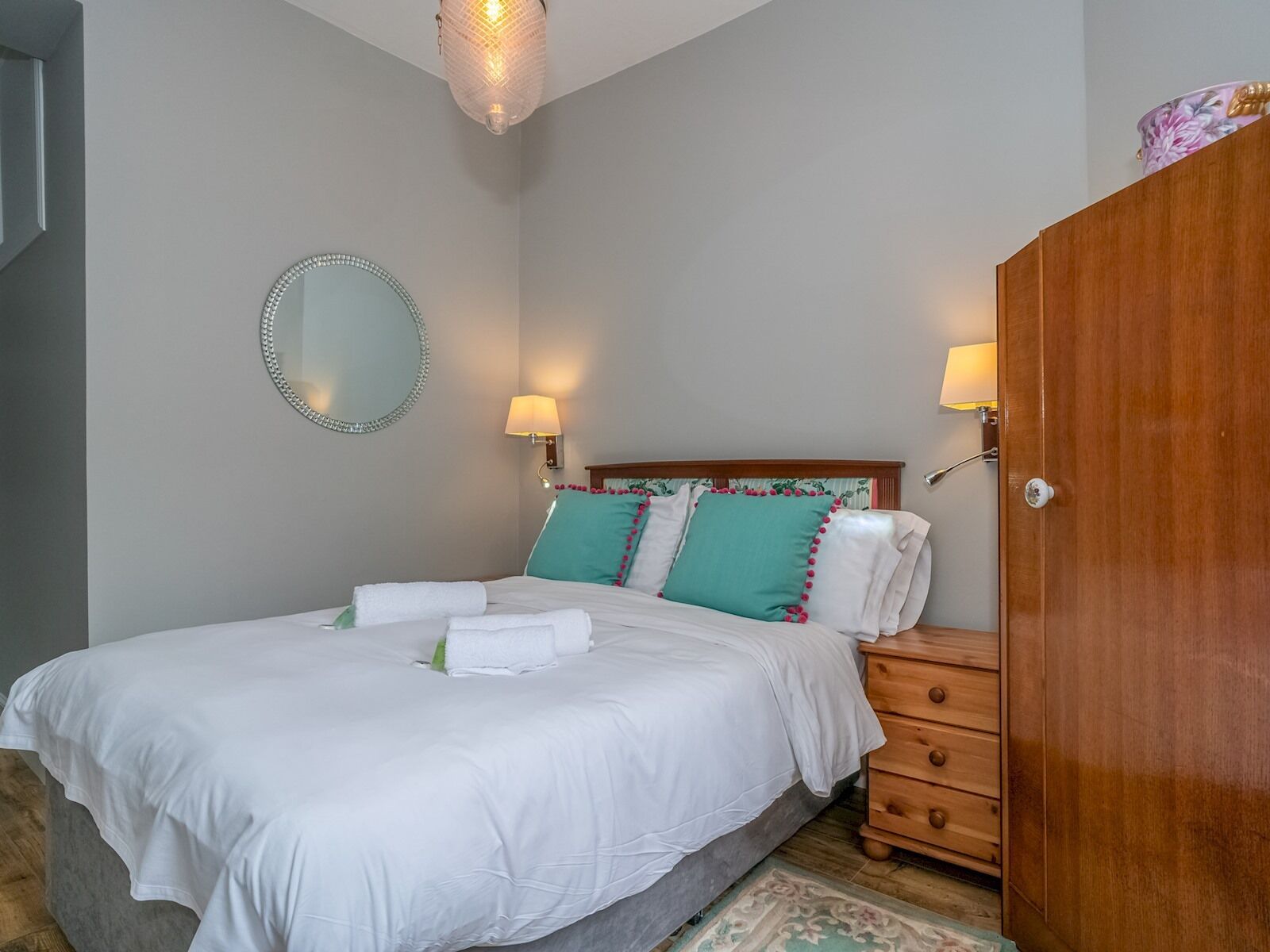 Una cama bien hecha con sábanas blancas y almohadas turquesa, un armario de madera y un espejo redondo en la pared.