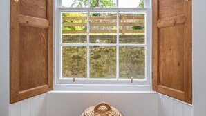 Una habitación con un gran ventanal con contraventanas de madera, vistas a un jardín con cercado de madera y una cesta tejida en el alféizar.
