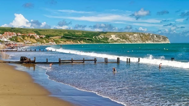 A coastal beach with waves, a wooden jetty, and a town in the background. - Dizzy Lows Sea View Apartment Swanage Town & Beach (Swanage)